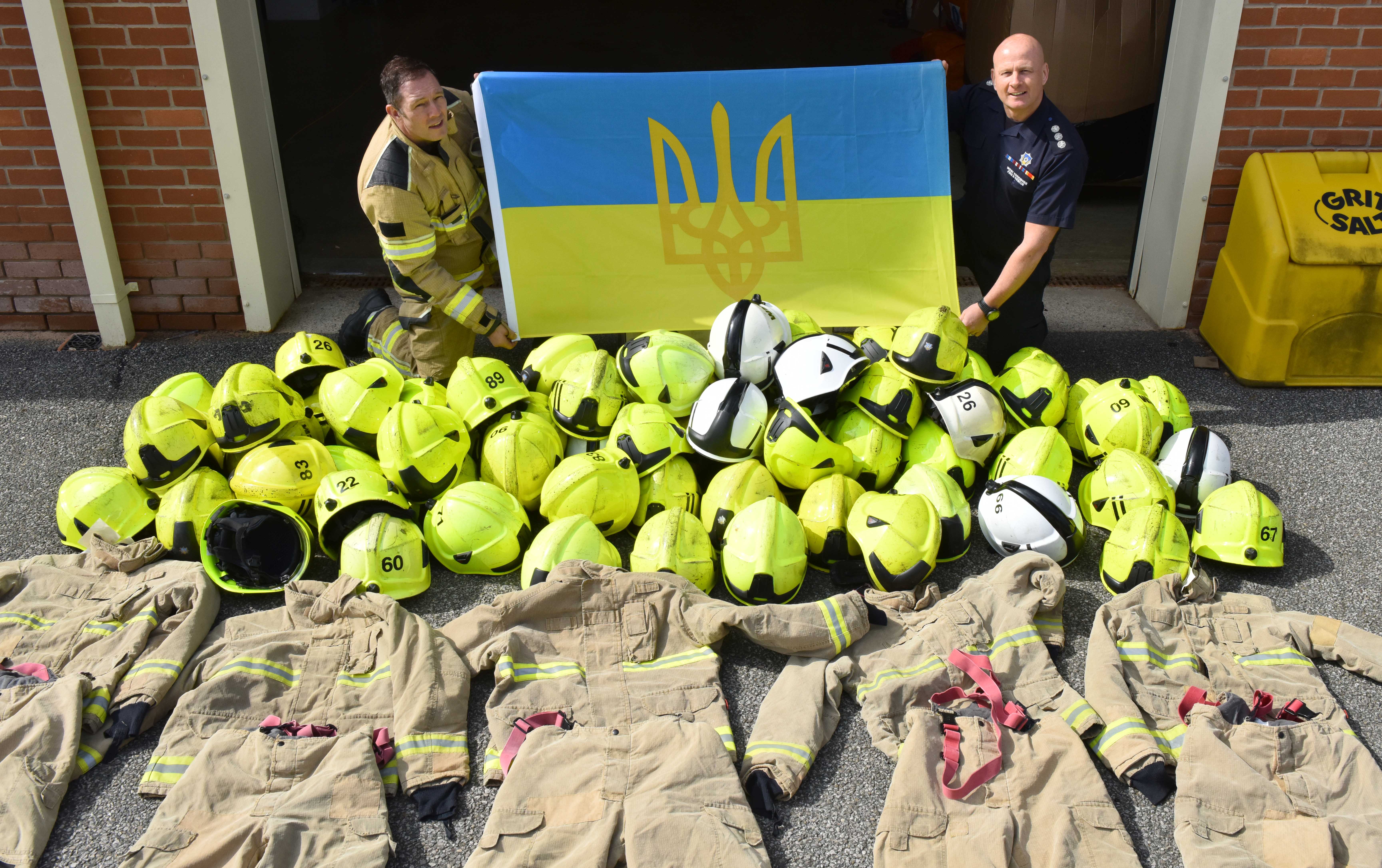 Helmets and Firefighter kit laid on the floor with 2 firefighters behind holding Ukrainian flag