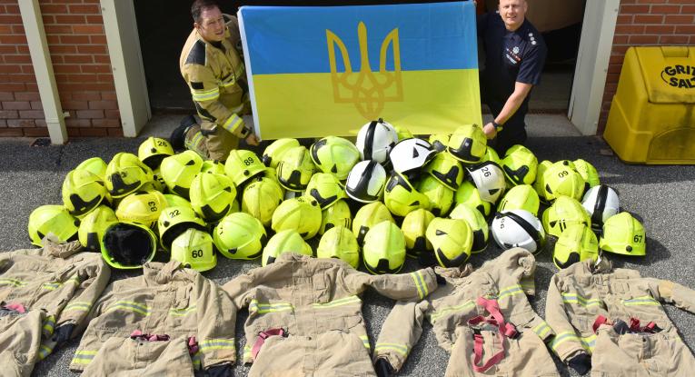 Helmets and Firefighter kit laid on the floor with 2 firefighters behind holding Ukrainian flag